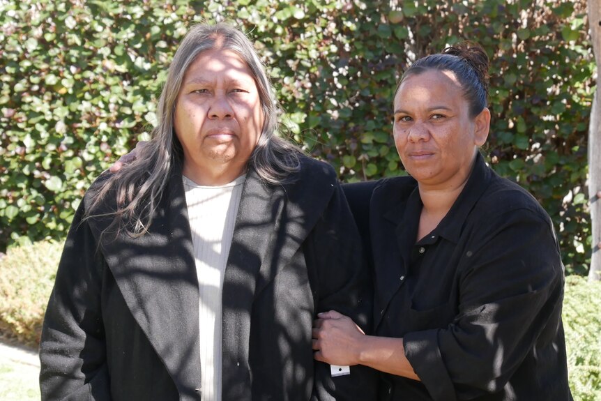 Two women, one in a black jacket and one in a black shirt, in front of a leafy green bush.