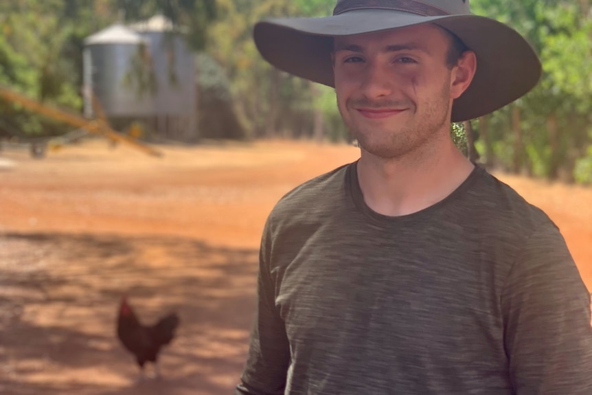 Smiling young man stands on a dirt road next to a chicken, wears a hat, brown tee, red earth behind, a hen, two water towers.