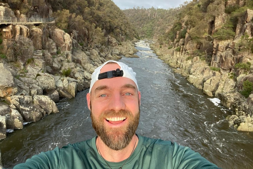 A man in a backwards cap smiles in front of a gorge in Tasmania