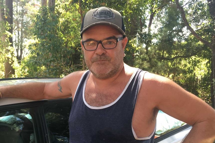 A man in a singlet and cap standing in front of his car.