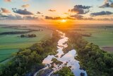 A photograph of a river that is running through farm fields and into a glowing sunrise on the horizon