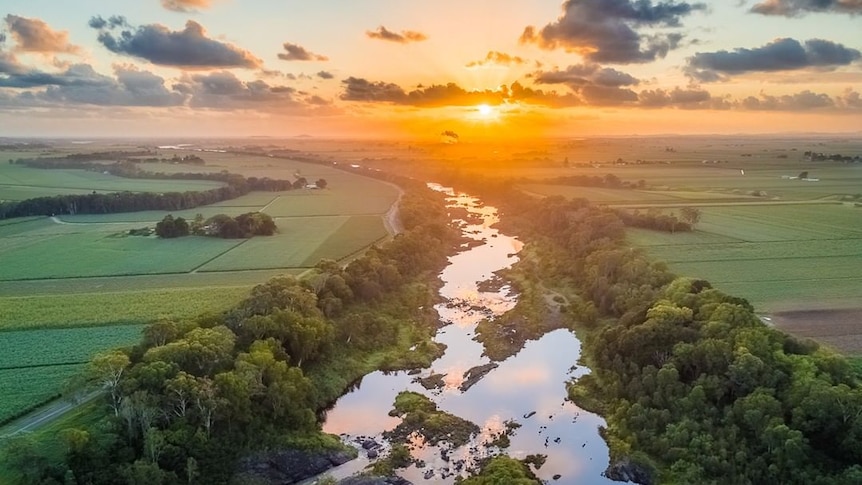 A photograph of a river that is running through farm fields and into a glowing sunrise on the horizon
