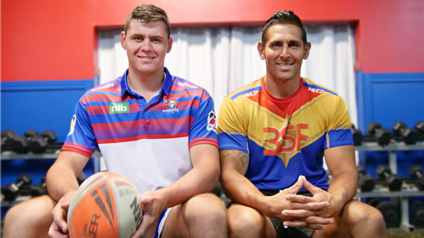 two men sitting on a bench in a gym smiling at the camera, one is holding a rugby ball