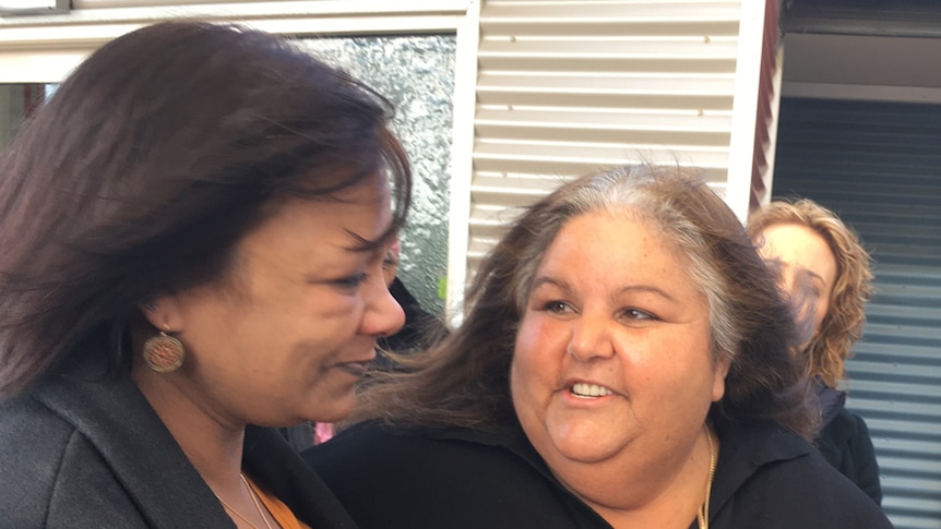 An indigenous woman startes contemplatively, almost crying, into the space in front of her while another woman looks proudly.