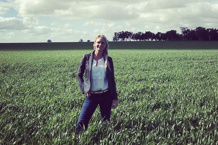 A woman is standing in the middle of a green field looking at the camera with a blue sky and clouds above.