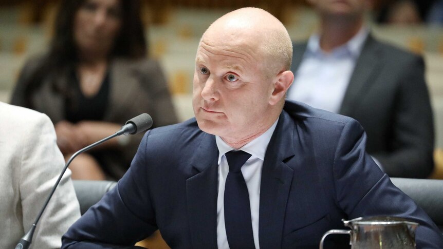Ian Narev, a bald man wearing a dark suit and tie, listens intently in his seat. There is a microphone in front of his face.