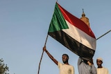 Three men stand on a car holding Sudanese flags in front of a mosque.