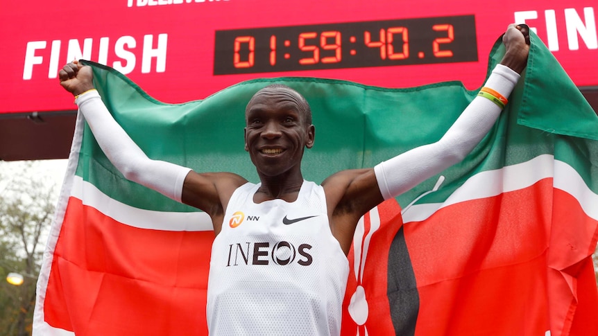 A Kenyan athlete stands holding his nation's flag behind his head as a clock displays his time for a world record.