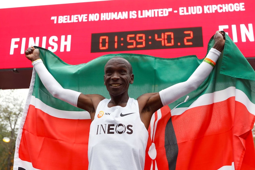 A Kenyan athlete stands holding his nation's flag behind his head as a clock displays his time for a world record.