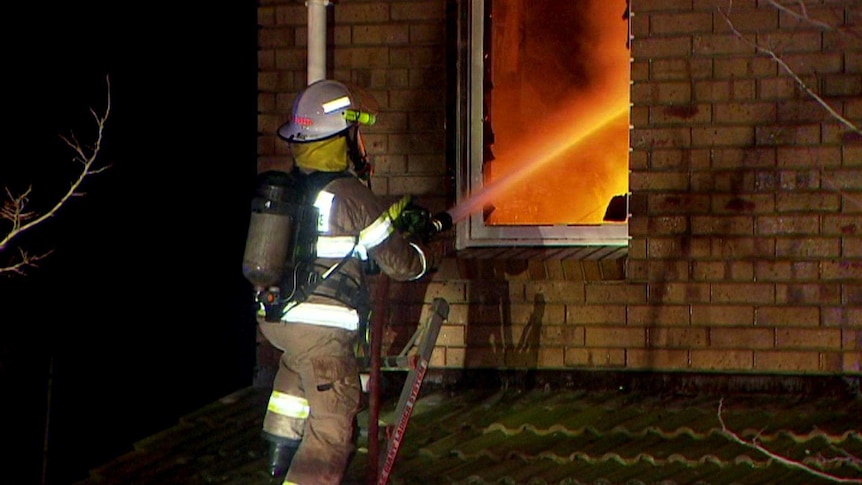 A firefighter on a ladder directs a water hose into broken second-storey window, with the room inside illuminated by flames.