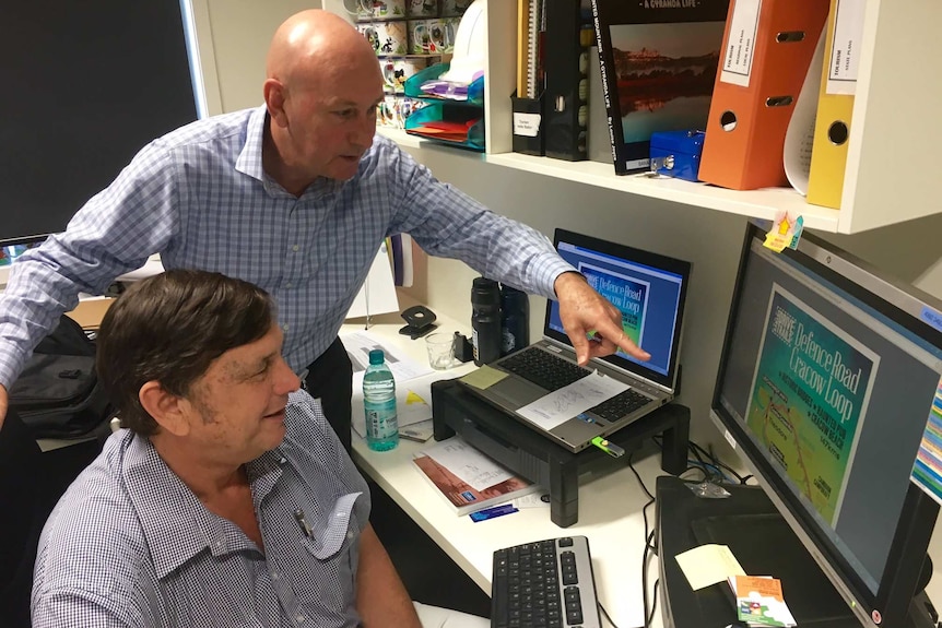 Two men in a cluttered council office, looking at a computer screen displaying a flyer for the Defence Road Crackow Loop tour.