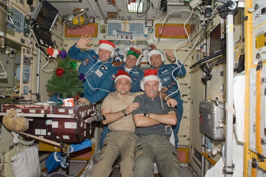 Five men wearing blue shirts and tan trousers floating in a space station and wearing santa hats