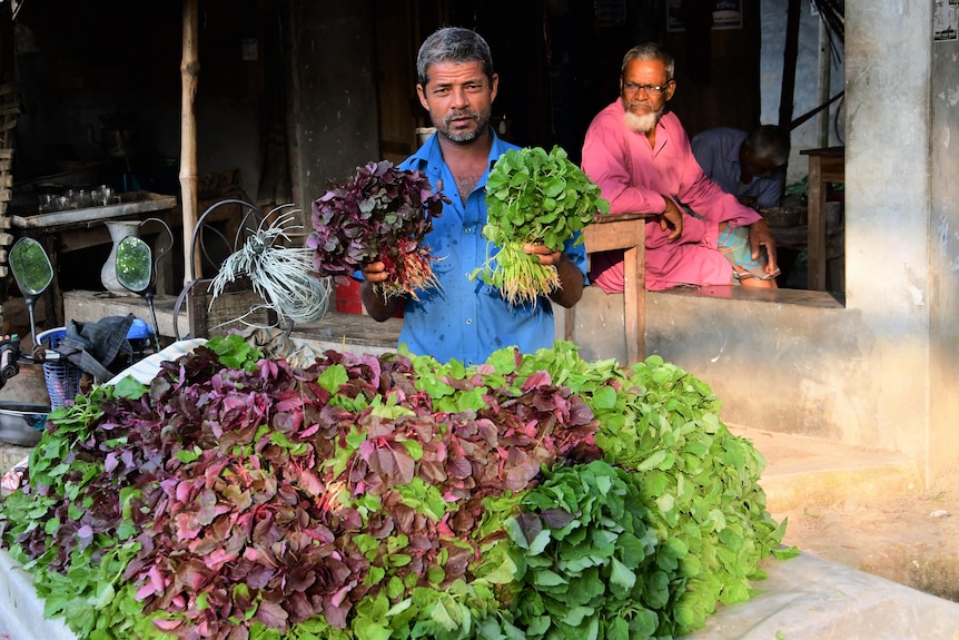 A farmer stands holding two bunches of leafy vegetables in front of a table piled with more.