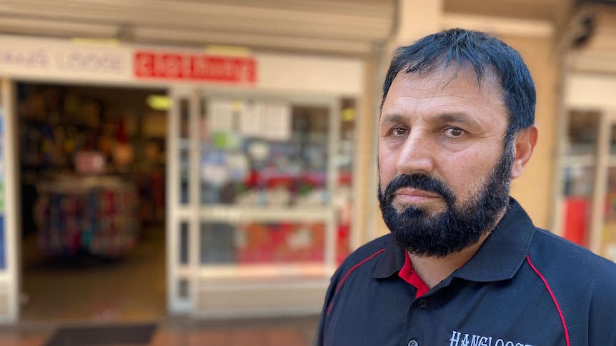 A bearded man in a black polo shirt stands in front of a shop, with a sad expression.