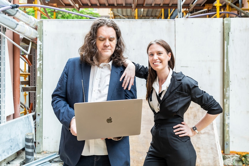 A man and a woman in dark suits stand in front of a laptop