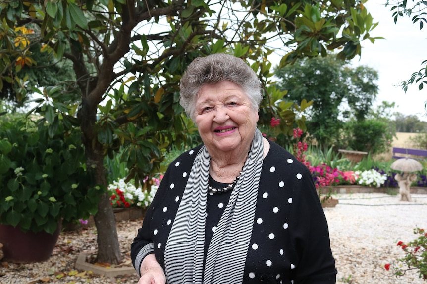 An elderly woman in a black shirt, standing in a garden.