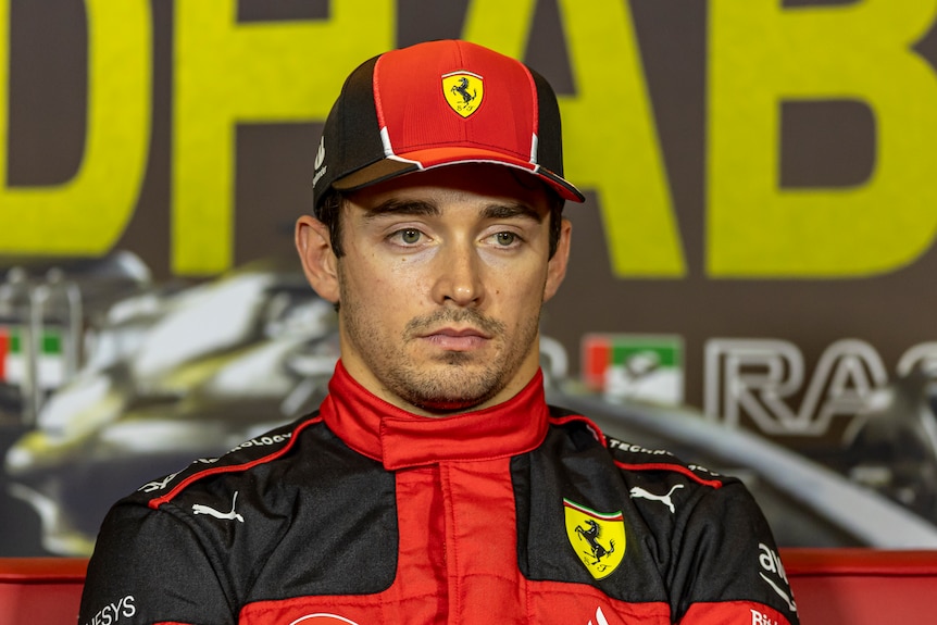 A driver in a Ferrari F1 cap and red shirt, looks on with an expressionless face