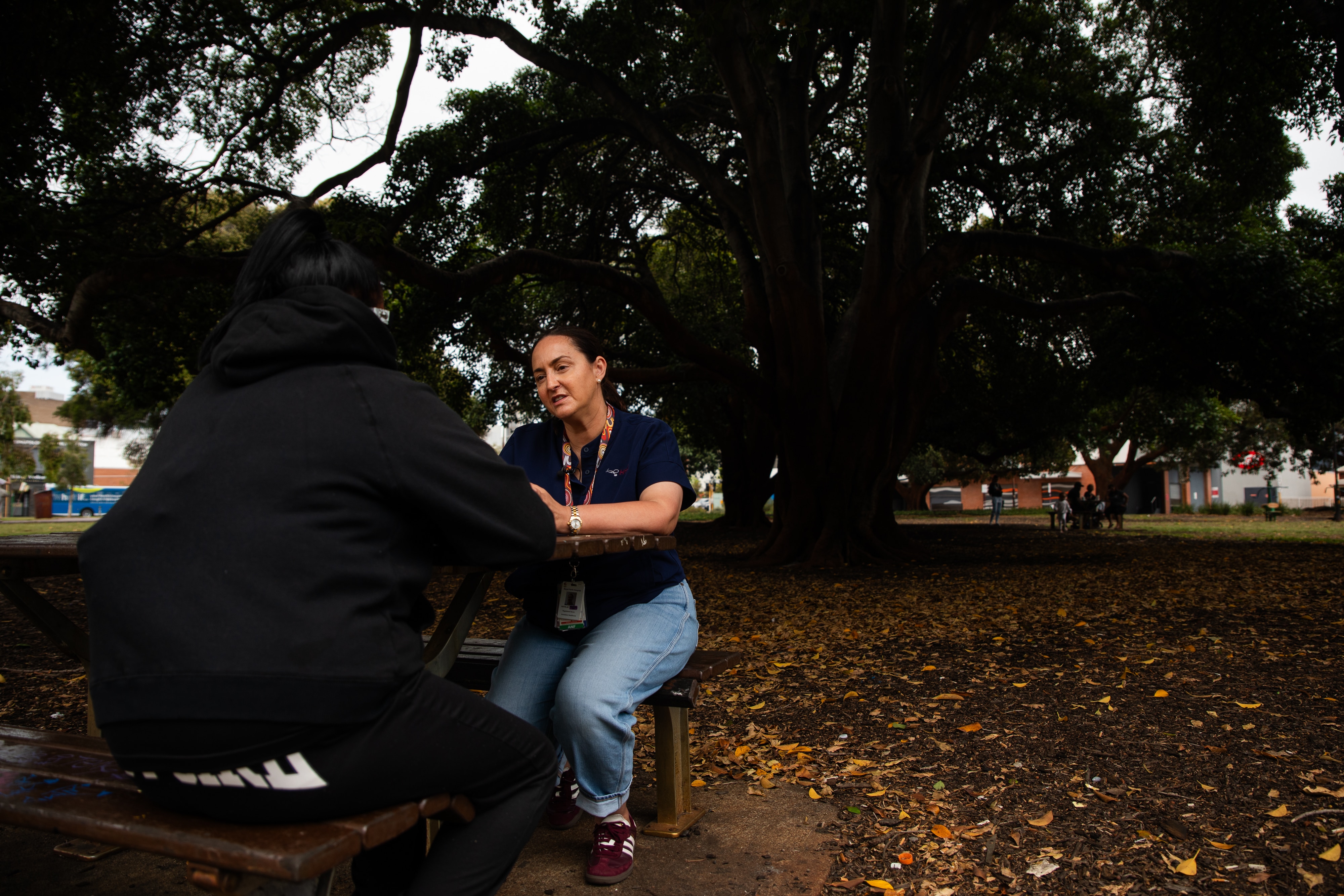 Duas mulheres sentadas a uma mesa no parque, uma delas de costas para a câmera.