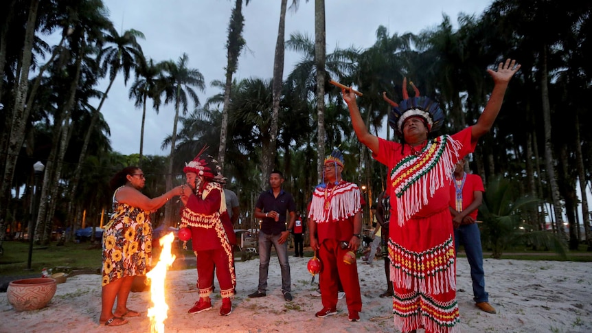 Members of Suriname indigenous tribes hold their arms towards the sky as a camp fire burns beside them.