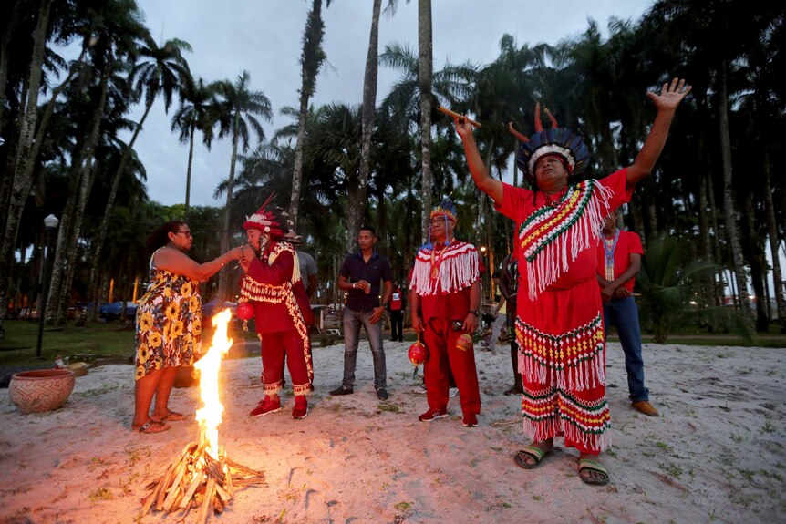 Members of Suriname indigenous tribes hold their arms towards the sky as a camp fire burns beside them.
