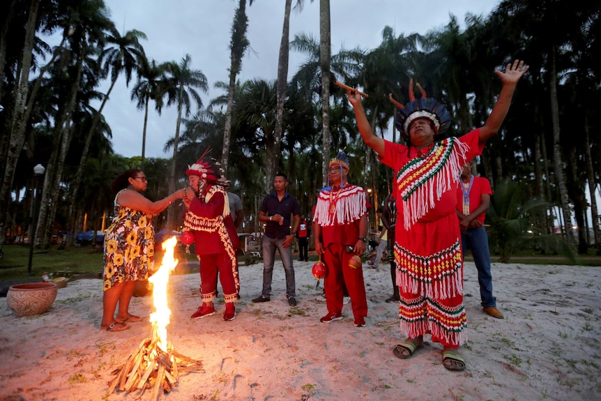 Members of Suriname indigenous tribes hold their arms towards the sky as a camp fire burns beside them.