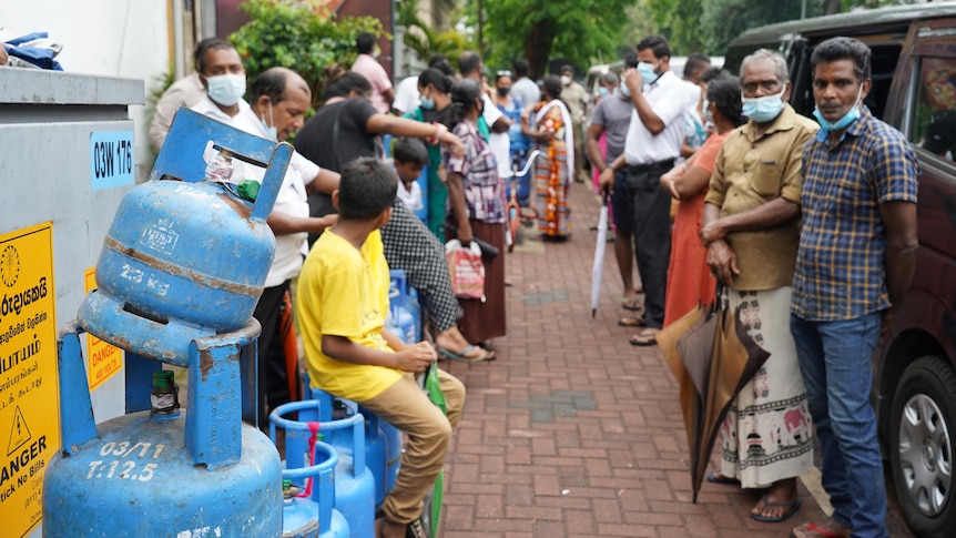 People stand and sit in a queue stretching down a footpath, some have brought gas bottles to be refilled