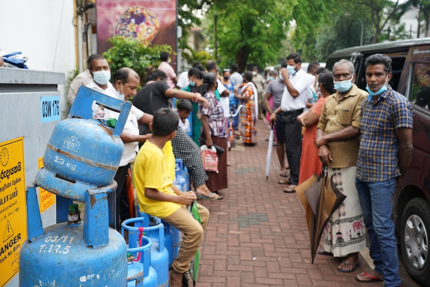 People stand and sit in a queue stretching down a footpath, some have brought gas bottles to be refilled