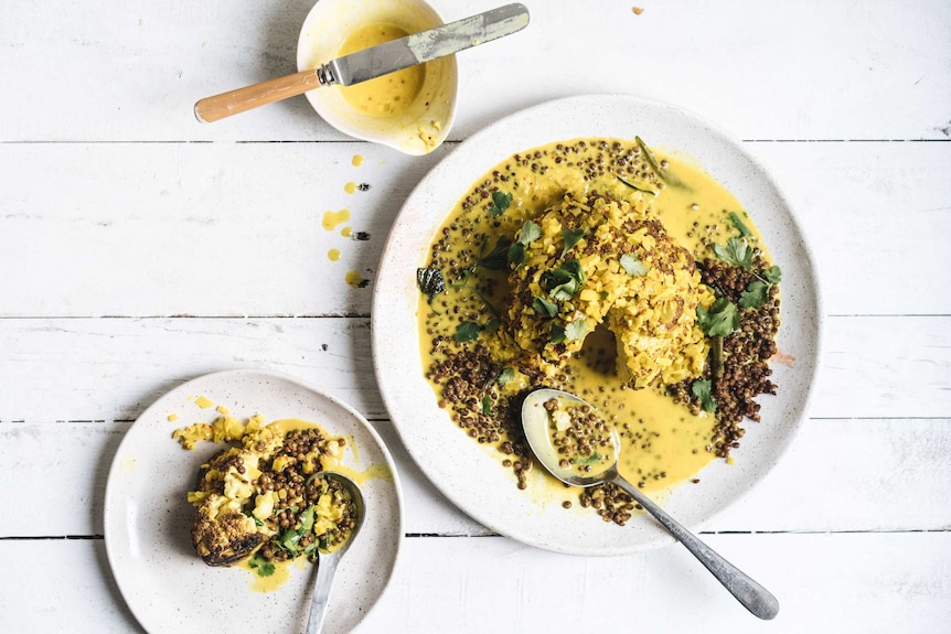 A small plate of roast cauliflower, tumeric sauce and lentils sits beside the main dish and extra sauce, for a vegetarian dinner