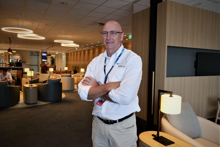 Man wearing casual business attire and lanyard stands in a lounge, arms folded.