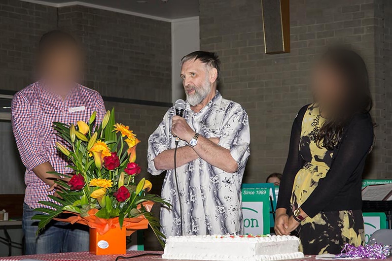 A man speaks into a microphone. He is flanked by two people whose faces are blurred.