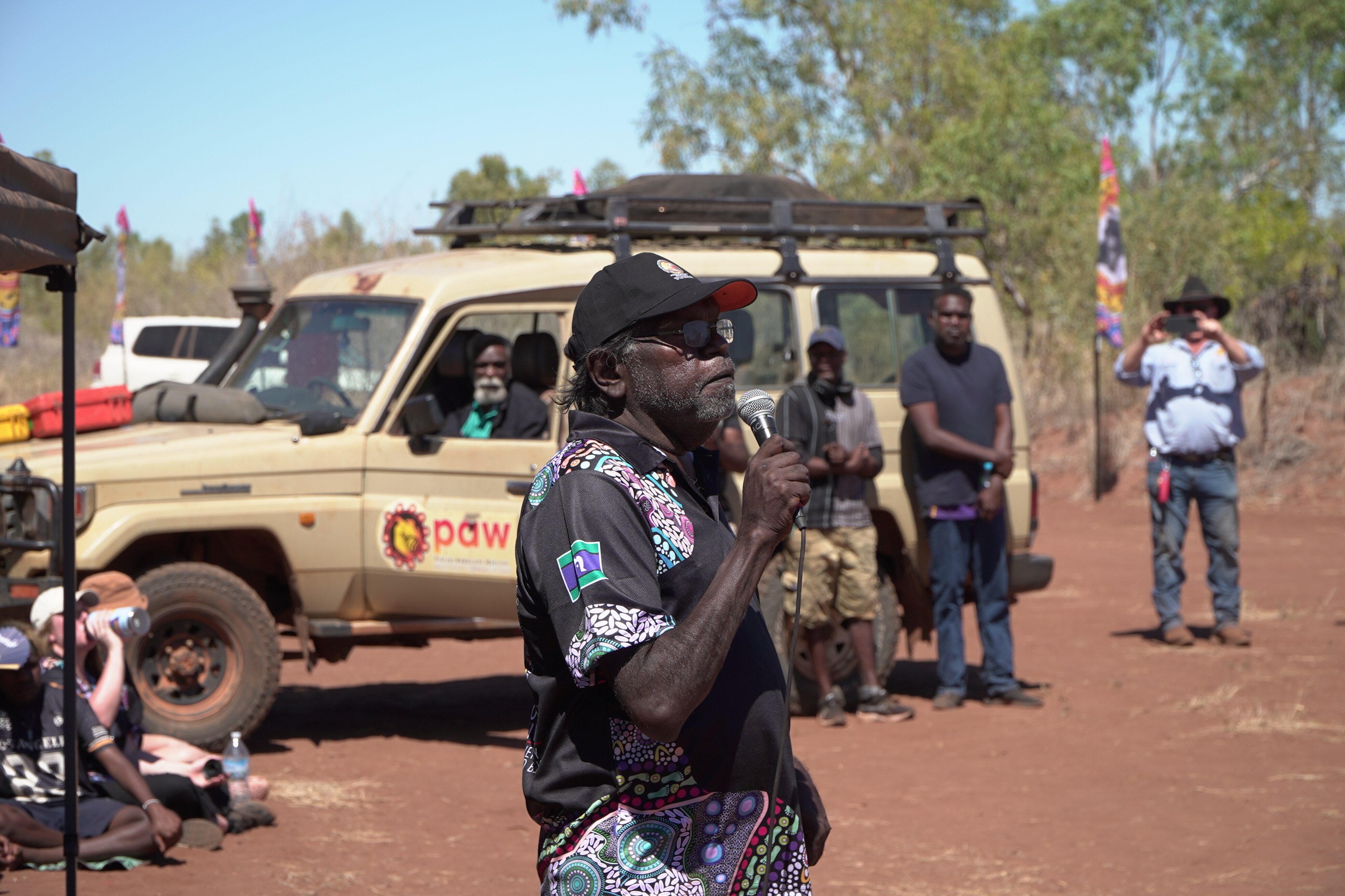 An Indigenous man speaking into a microphone as others watch on.