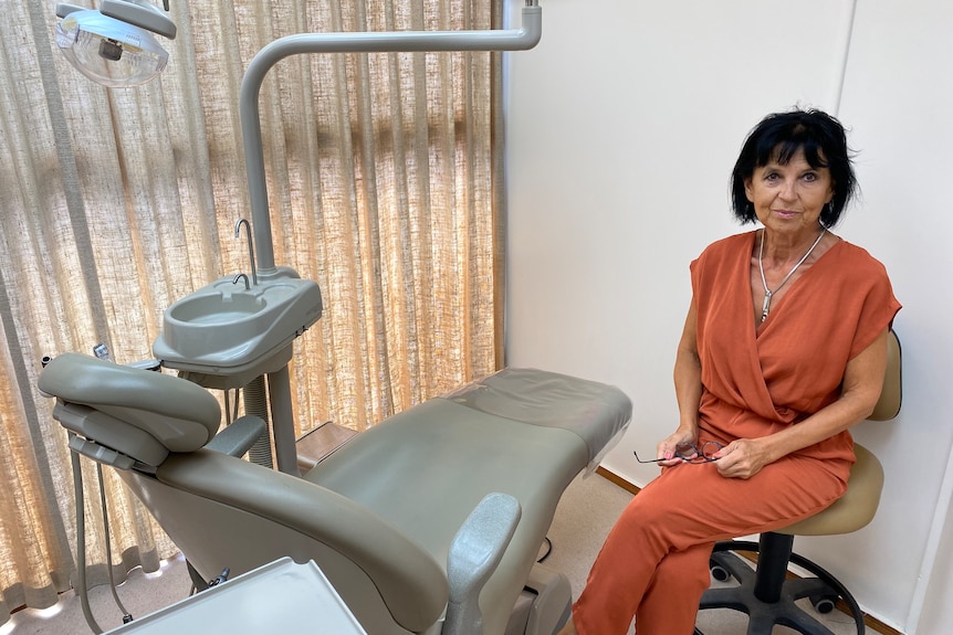 a woman with dark short black hair wears an orange jumpsuit and sits aside a dental chair