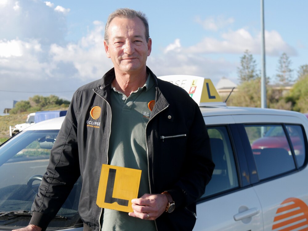 A man smiling holding an L-plate sign standing in front of a white car