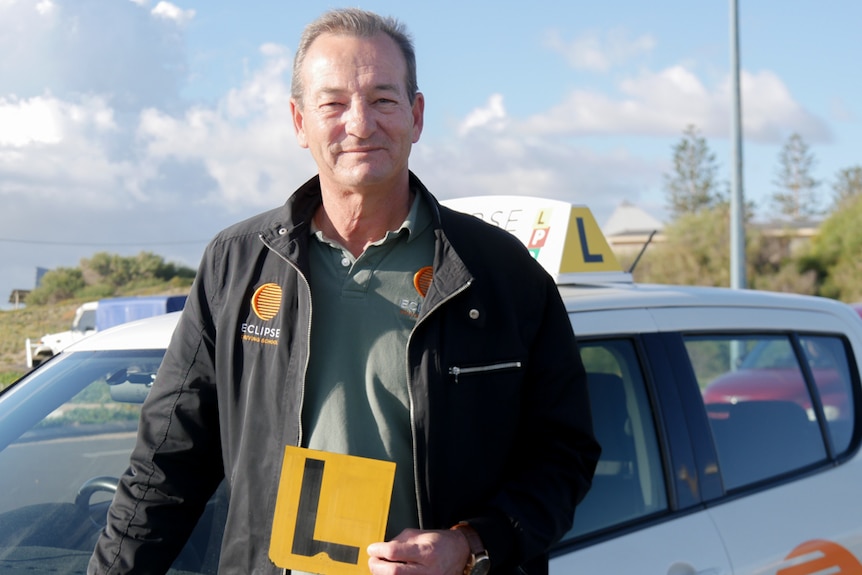 A man smiling holding an L-plate sign standing in front of a white car