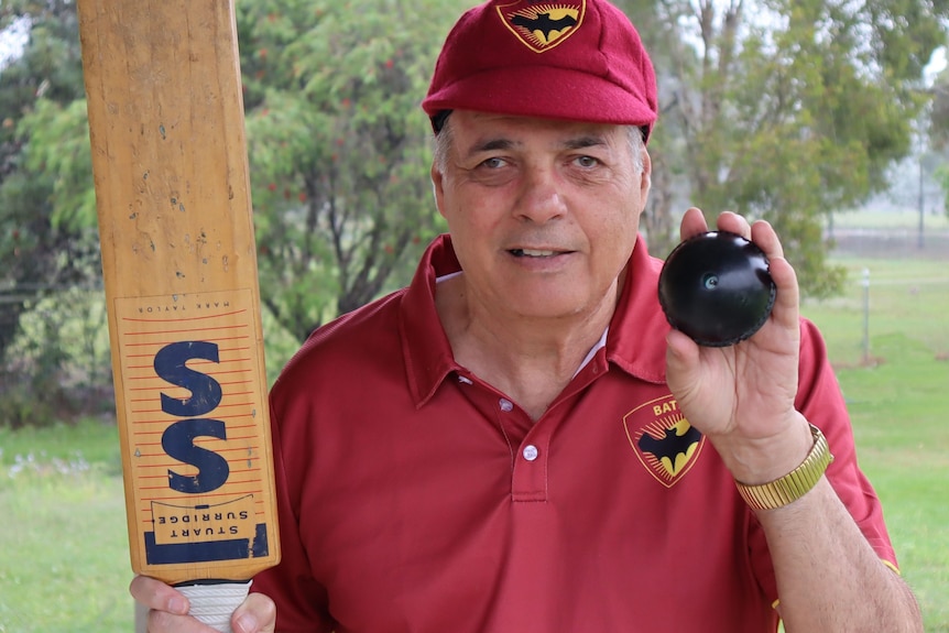Man in red shirt and cap holding cricket bat and black ball