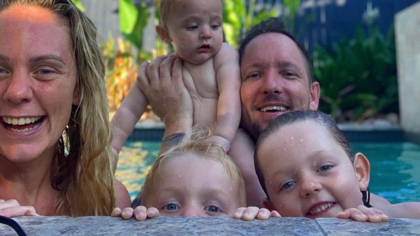 A blonde woman smiles while sitting in the pool with her husband and three small children.