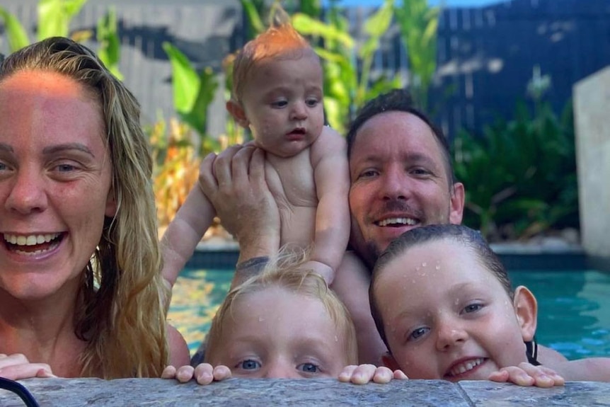 A blonde woman is smiling while sitting in the pool with her husband and three children. 