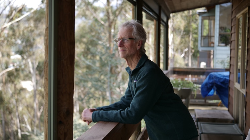 A white-haired man wearing glasses leans on a balcony railing surrounded by bushland.