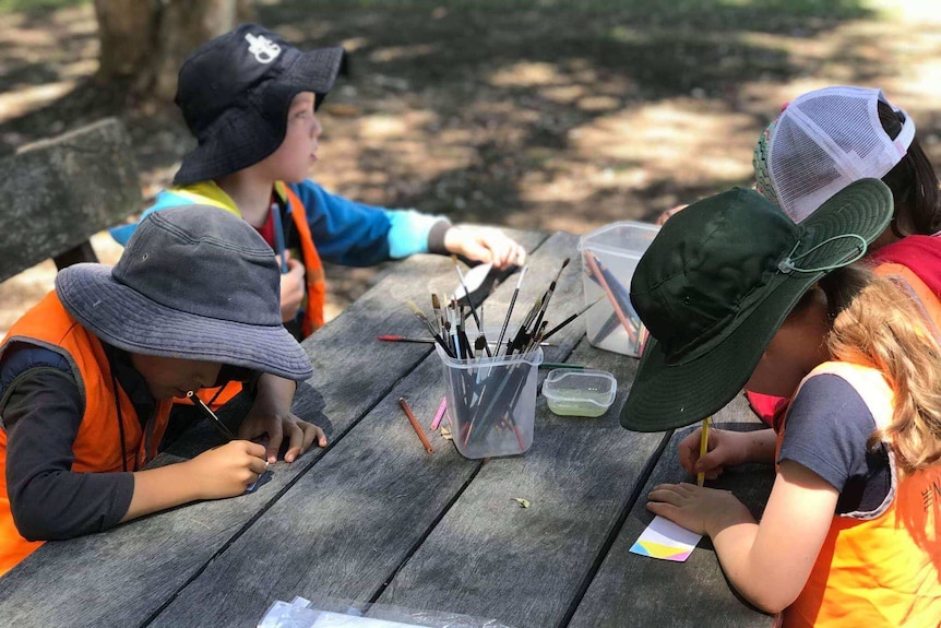 Four children, wearing wide-brimmed hats sitting at a wooden table outside, drawing.