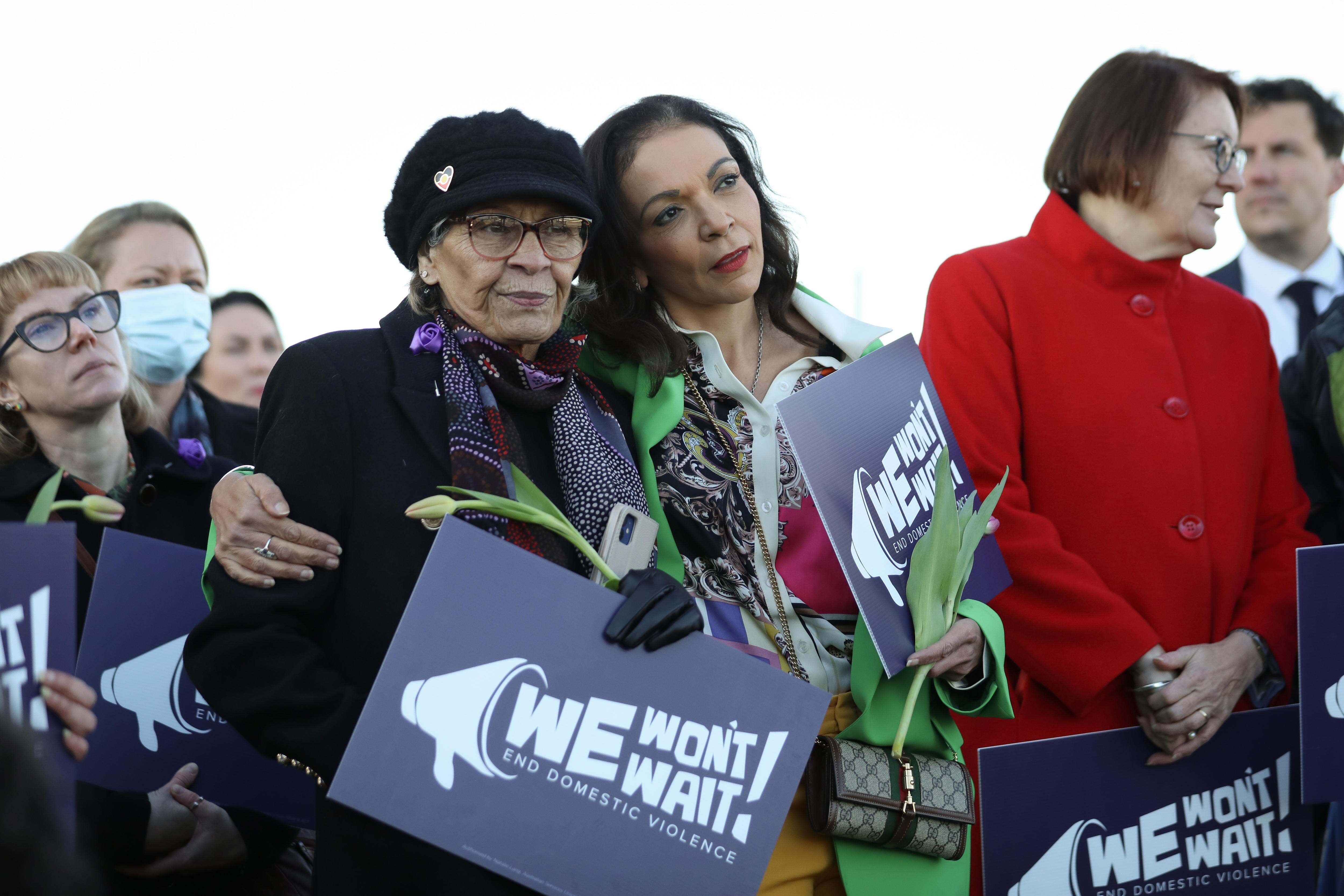 Aly puts her arm around a woman while they hold signs and flowers outdoors.