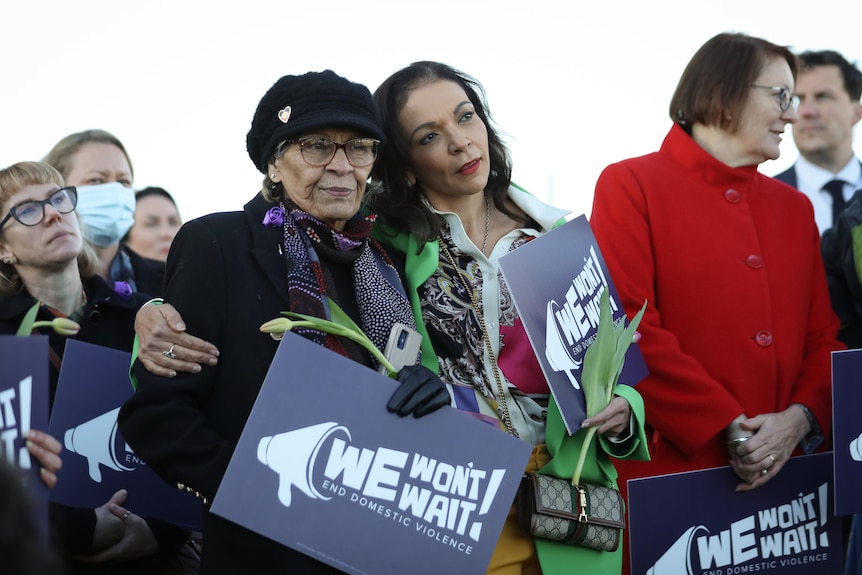 Aly puts her arm around a woman while they hold signs and flowers outdoors.