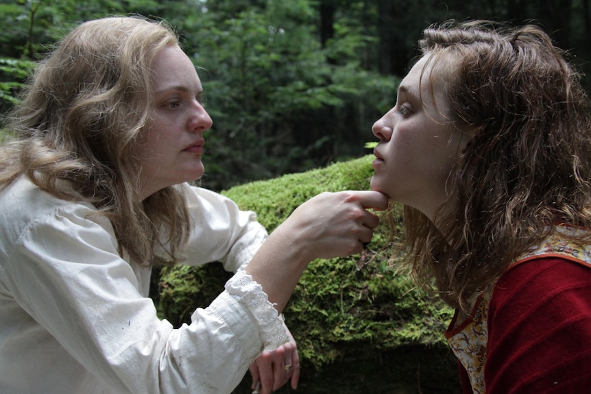 Two women face each other in forest, blonde woman on left lifts chin of uneasy looking dark haired woman with her hand.
