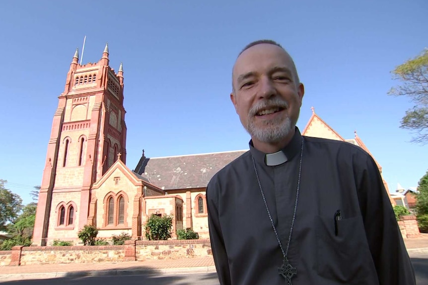 A man in a black shirt with a priest's collar stands in front of the St Andrew's church in Adelaide.