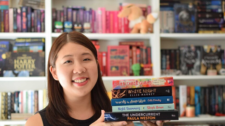 A young Asian woman stands in a room filled with books, balancing a stack of books