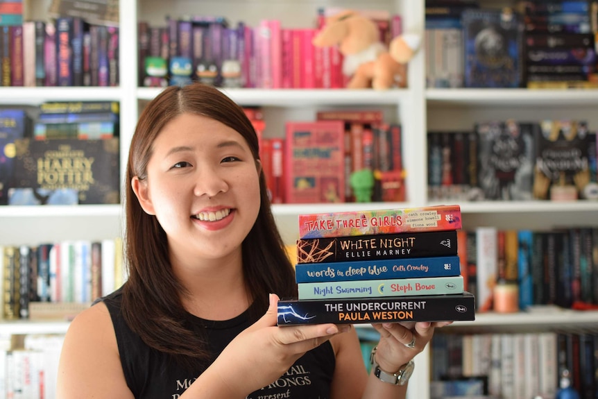 A young Asian woman stands in a room filled with books, balancing a stack of books