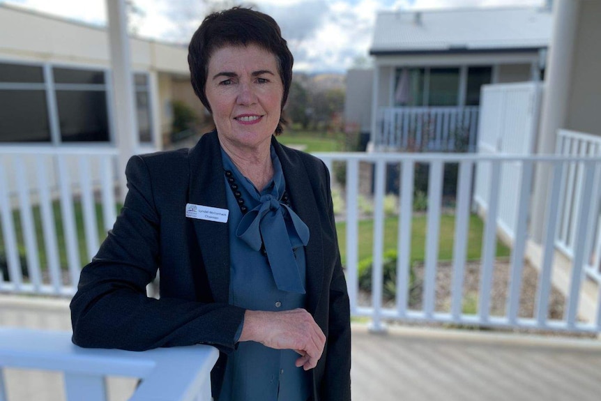 A woman with short, dark hair, wearing a blue top and black blazer, stands on a verandah.