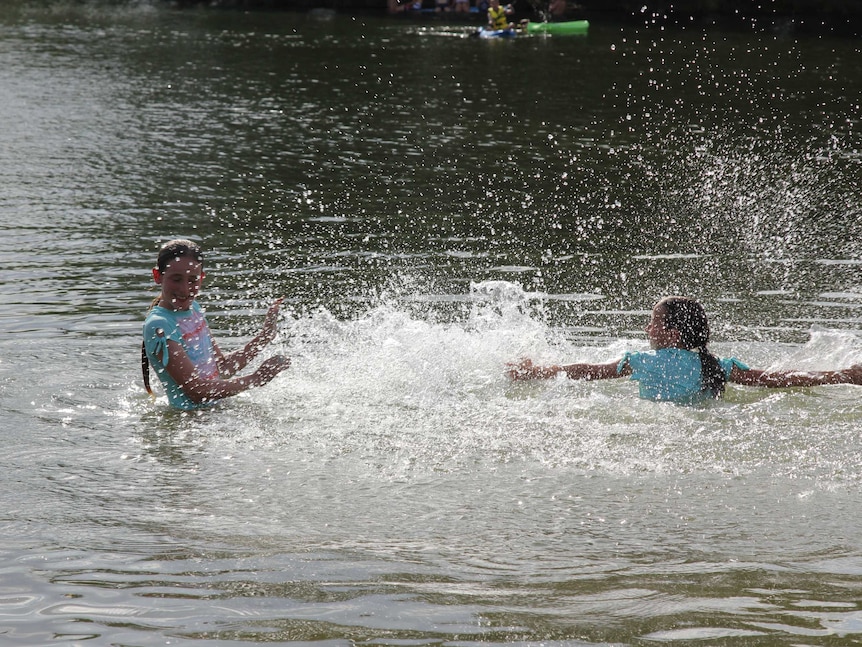 Two girls splash in water while people kayak behind them.
