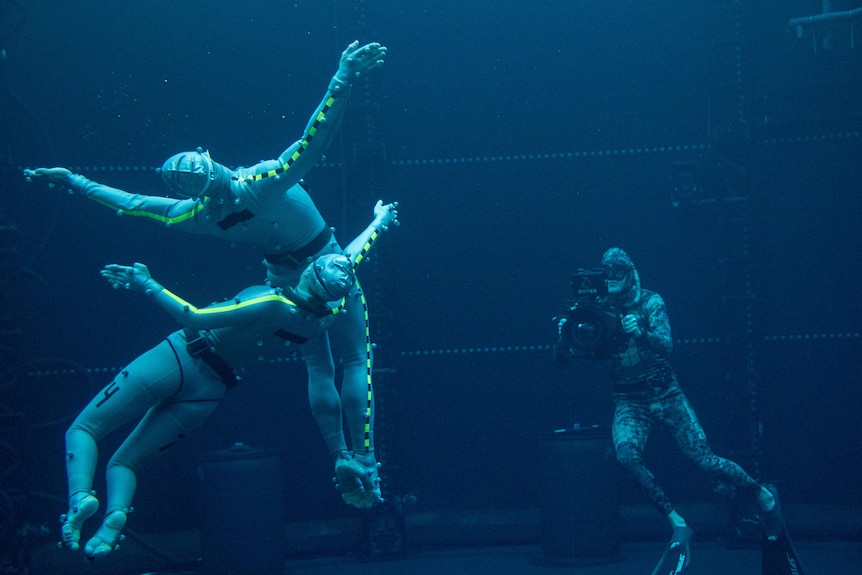 actors underwater swim close together in cgi wetsuits, with film operator with camera in background