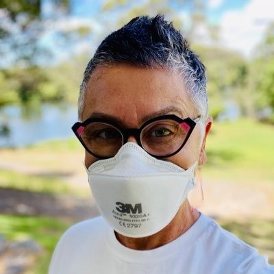 A woman standing outside in a white t-shirt with grey, cropped hair and wearing a medical mask.
