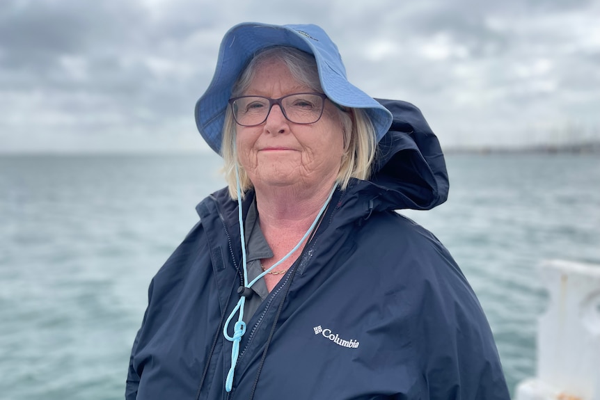 A woman in a blue hat and wearing a blue jacket is by the ocean. She has a serious expression.